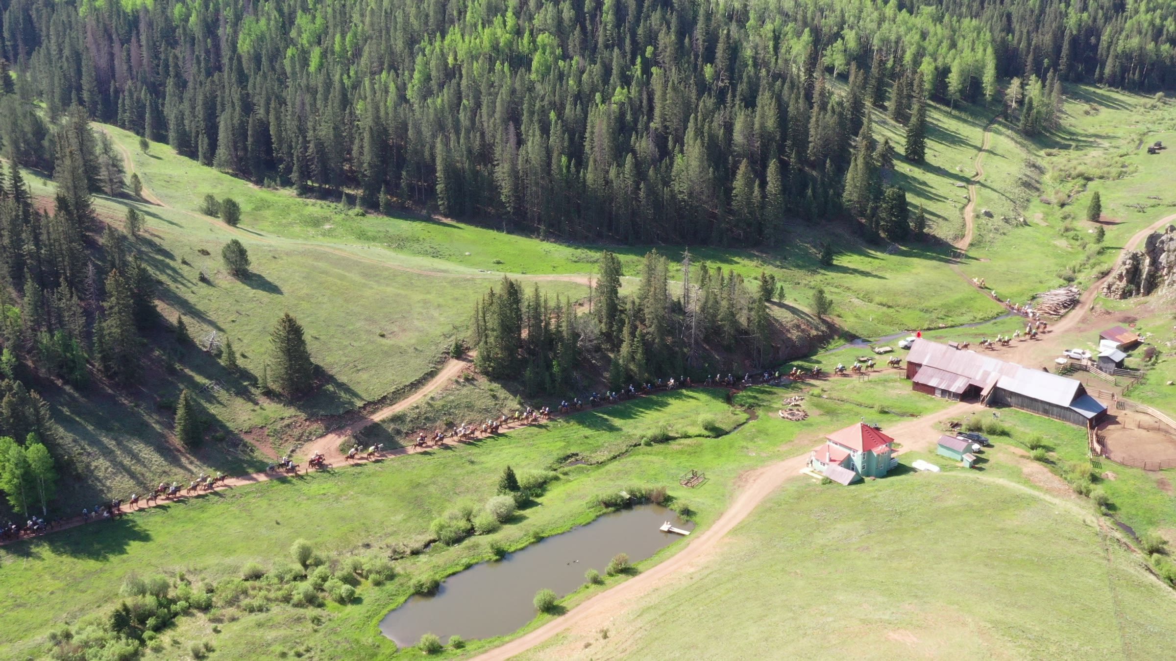 Aerial view of the weathered Elk Glade barn and red-roofed ranch house above the pond, a line of riders following the creek below spruce slopes
